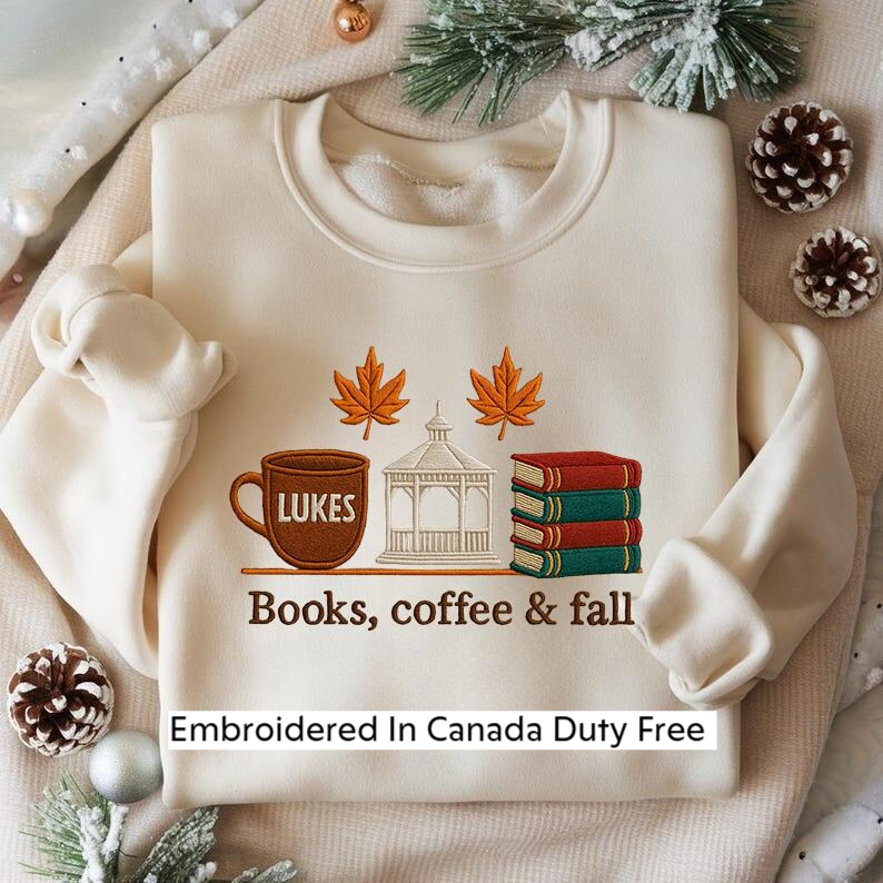 A white sweater with a graphic design on it, featuring a cup of coffee, a stack of books, and autumn leaves, along with the text "Books, coffee & fall" and "Embroidered In Canada Duty Free".