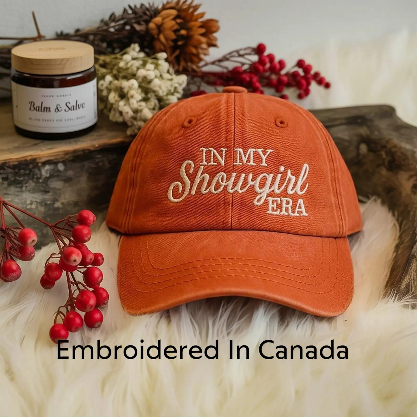 An orange baseball cap with the text "IN MY SHOWGIRL ERA" embroidered on it, placed on a white fur surface. There is also a jar of balm and some red berries in the background.