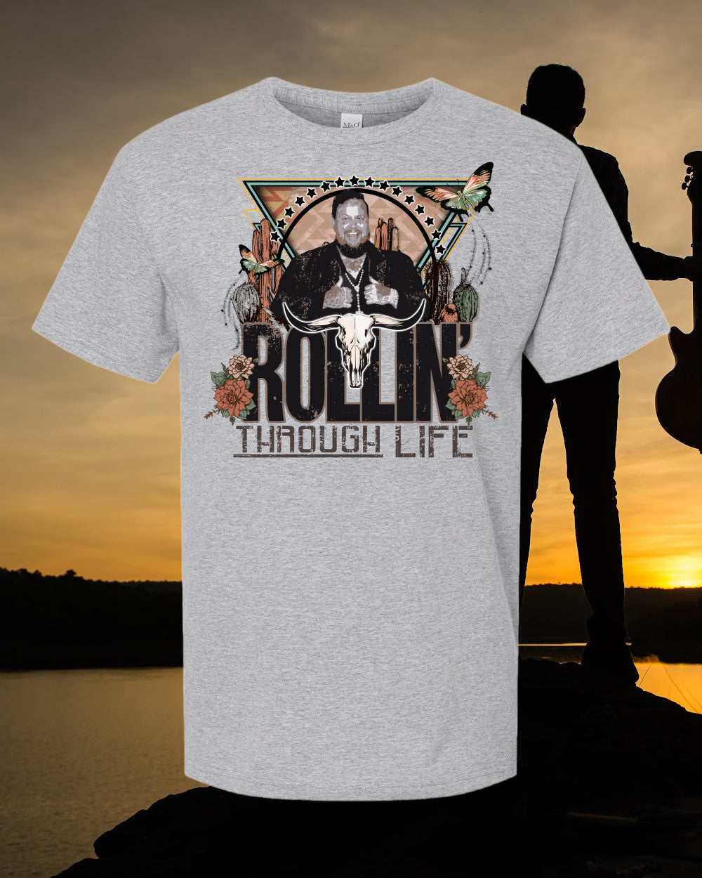 A graphic t-shirt with a design that includes text 'ROLLIN' THROUGH LIFE' and imagery of a skull, butterflies, and floral elements against a sunset background.
