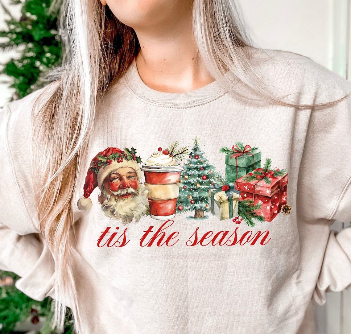 A sweatshirt with a Christmas themed transfer design featuring Santa Claus, a Christmas tree, and the text 'Tis the Season'.