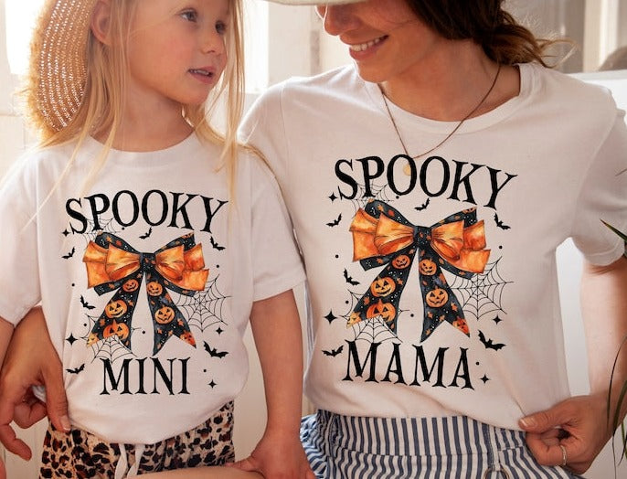A child and an adult wearing white t-shirts with the text 'Spooky Mama' and 'Spooky Mini' on them, featuring a bow and pumpkin graphics.