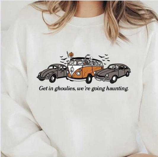 A white sweatshirt with a graphic transfer featuring a cartoonish design of a van and a car, and the text 'GET IN GHOULS. WE'RE GOING HAUNTING.