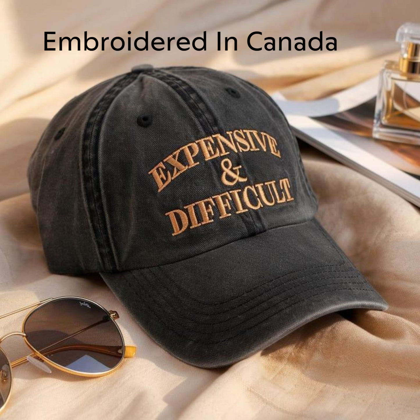 a black baseball cap with the text "EXPENSIVE & DIFFICULT" embroidered on it, placed on a beige fabric surface. There are also sunglasses and a perfume bottle visible in the background.