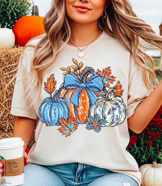 Woman wearing a t-shirt with pumpkins and leaves design, sitting on hay bales with pumpkins and flowers in the background.