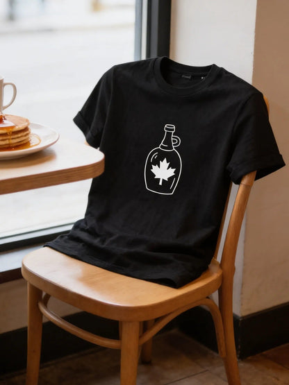 A black t-shirt with a maple leaf design is draped over a wooden chair, with a plate of pancakes and a cup of coffee on a table in front of it.