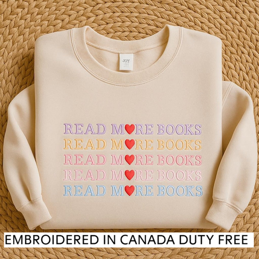 A beige sweater with the text "READ MORE BOOKS" embroidered on it, along with a tag that says "JOY". The sweater is displayed on a woven mat, and there is text at the bottom of the image that says "EMBROIDED IN CANADA DUTY FREE".