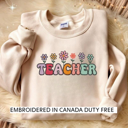 A beige sweater with a colorful embroidered design that says "TEACHER" in a playful font, surrounded by flower-like illustrations.