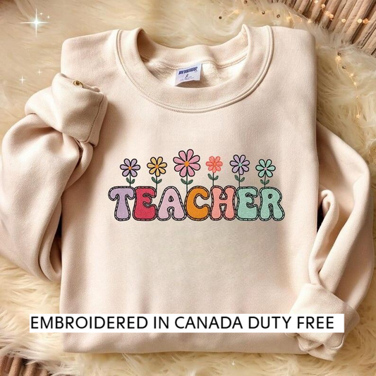 A beige sweater with a colorful embroidered design that says "TEACHER" in a playful font, surrounded by flower-like illustrations.