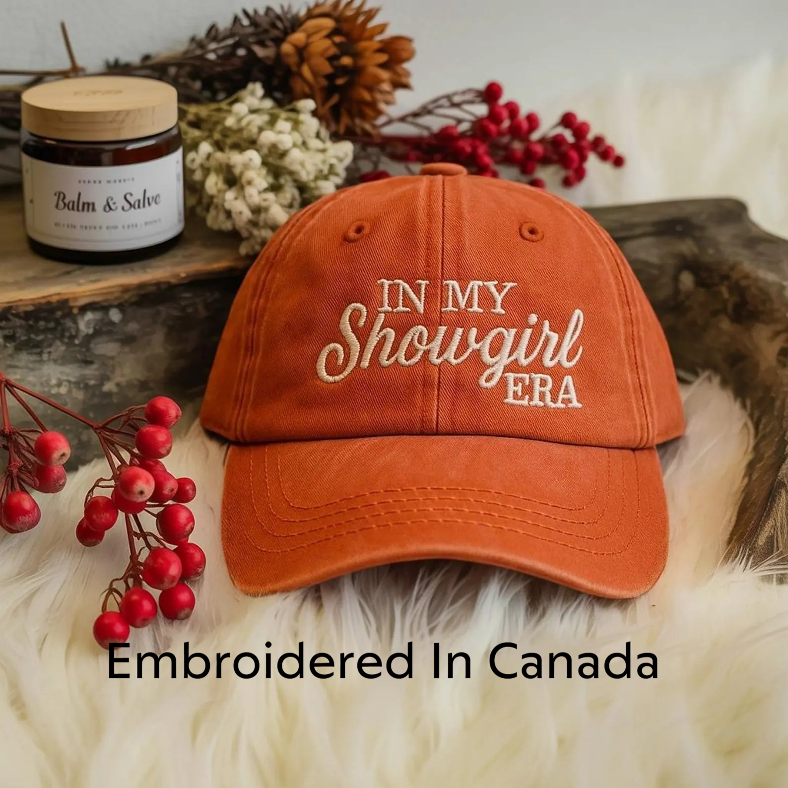 An orange baseball cap with the text "IN MY SHOWGIRL ERA" embroidered on it, placed on a white fur surface. There is also a jar of balm and some red berries in the background.