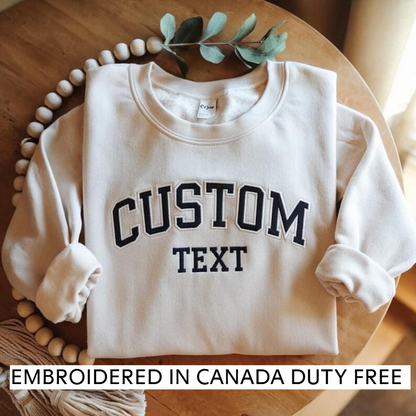 A white sweatshirt with the text "CUSTOM TEXT" printed on it, along with a tag that says "Embroidered in Canada Duty Free". The sweatshirt is placed on a wooden surface, and there is a sprig of greenery nearby.