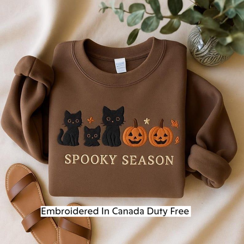 A brown sweater with a Halloween-themed design, including three black cats and two jack-o-lanterns, along with the text "SPOOKY SEASON" and "Embroidered In Canada Duty Free".