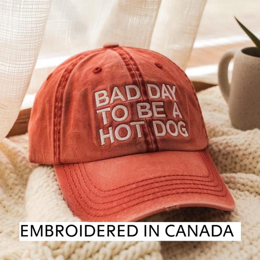 A red baseball cap with the text "BAD DAY TO BE A HOT DOG" embroidered on it, placed on a white blanket.