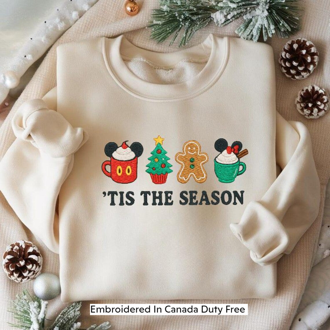A white sweater with a festive design featuring a Christmas tree, gingerbread man, and cupcakes, along with the text "TIS THE SEASON". The sweater is embroidered with the text "Embroidered In Canada Duty Free".