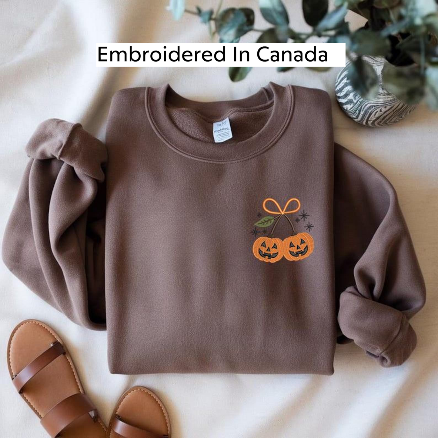 a brown sweater with a Halloween-themed design, including two jack-o-lanterns and a bow, laid out on a white surface. There is also a pair of sandals and a potted plant in the background.